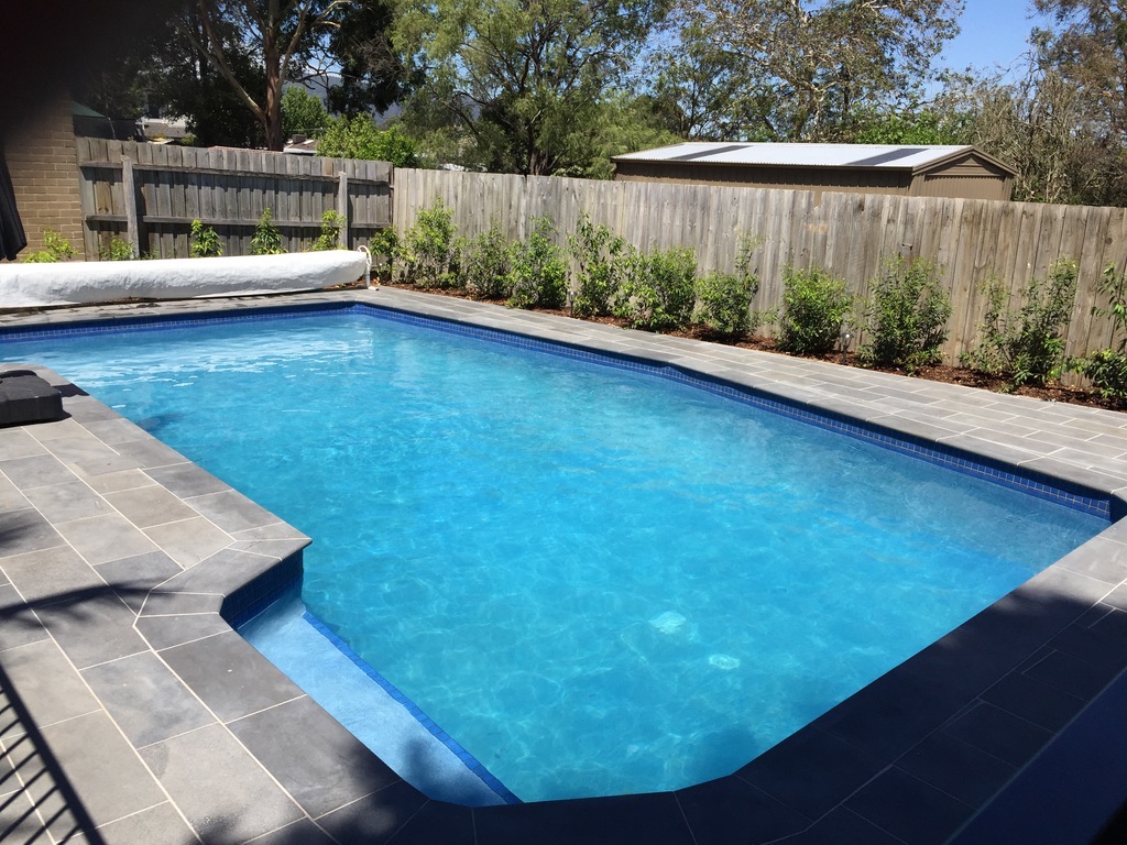 The image displays a well-maintained swimming pool situated in a serene outdoor environment. The pool features clear, sparkling water illuminated by natural sunlight, making it highly inviting. Surrounding the pool, there are neatly laid grey tiles that enhance the overall aesthetic. A light-coloured, partial cover is seen on the pool area, likely for maintenance or safety purposes. The background includes a wooden fence and lush greenery, providing a tranquil atmosphere. This image embodies the essence of a perfect outdoor leisure space, ideal for relaxation and enjoyment.