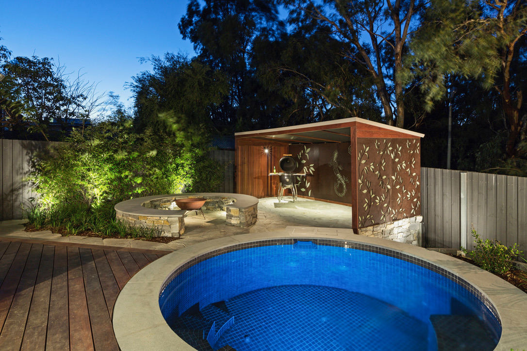 The image showcases a beautifully designed swimming pool area set amidst lush greenery, illuminated by soft landscape lighting at dusk. The pool features a circular shape with mosaic tiles, enhancing its appeal. Adjacent to the pool is a rustic-style wooden shelter, possibly serving as a relaxation space or outdoor entertainment area. The setting suggests a tranquil environment, ideal for leisure and gatherings. The incorporation of natural elements such as trees and shrubs adds warmth and a sense of privacy to the space, making it a perfect spot for swimming and outdoor activities.