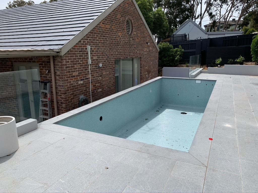 The image depicts an unfilled swimming pool positioned adjacent to a brick house. The pool has light blue walls and a smooth surface, with some debris visible at the bottom. The surrounding patio is paved and equipped with glass fencing, providing a modern touch. Nearby, the brick house features a slanted roof, and the overall environment appears tidy, with greenery in the background, indicating a well-maintained property. This is representative of both the installation and repair process commonly handled by swimming pool repair services.