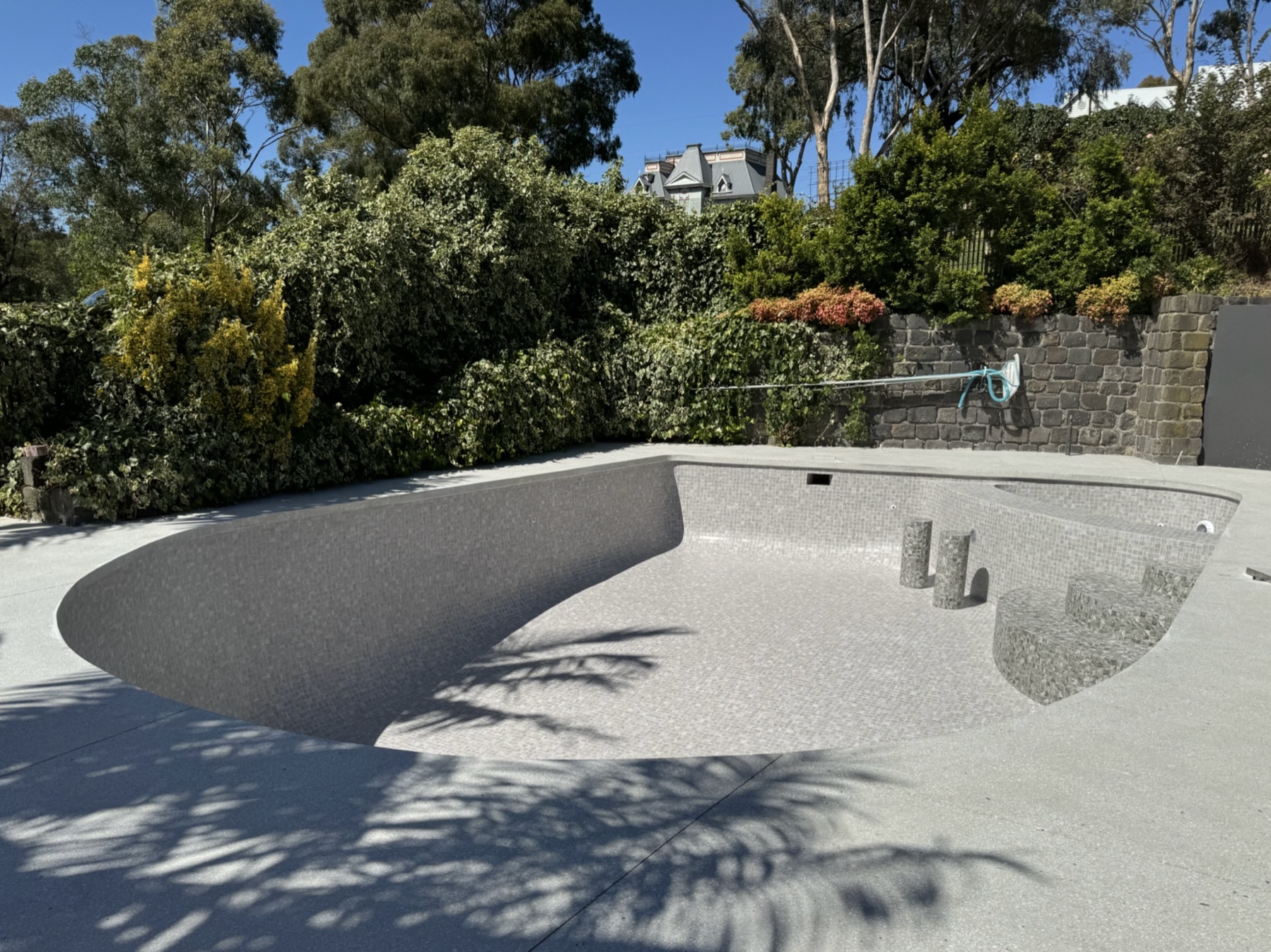 The image showcases an empty swimming pool situated in a well-maintained outdoor space. The pool features a modern design with a smooth, pebble-like surface that reflects the sunlight. Surrounding the pool are lush green plants and trees, suggesting a serene environment. The layout appears spacious, with ample decking around the pool area, indicating a focus on a clean, appealing aesthetic. The absence of water and any visible repairs hints at a current renovation or maintenance phase, emphasising the need for professional swimming pool repair services.