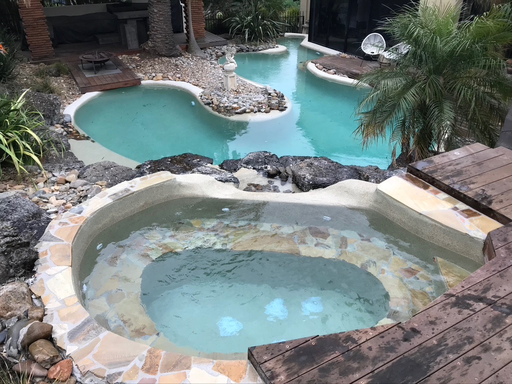 The image showcases a beautifully designed swimming pool area featuring a crystal-clear water pool with an adjoining spa. The pool is semi-circular with natural rock formations as a boundary, enhancing the natural aesthetic. Surrounding the pool, lush greenery, palm trees, and decorative rocks create a serene ambience. There are wooden deck areas adjacent to the pool, providing a space for relaxation and enjoyment. A small water feature with a statue can be seen in the distance, adding an elegant touch to the outdoor space. Altogether, it highlights a well-maintained pool area that exemplifies the quality of work Pools That Work provides in swimming pool repair and maintenance.