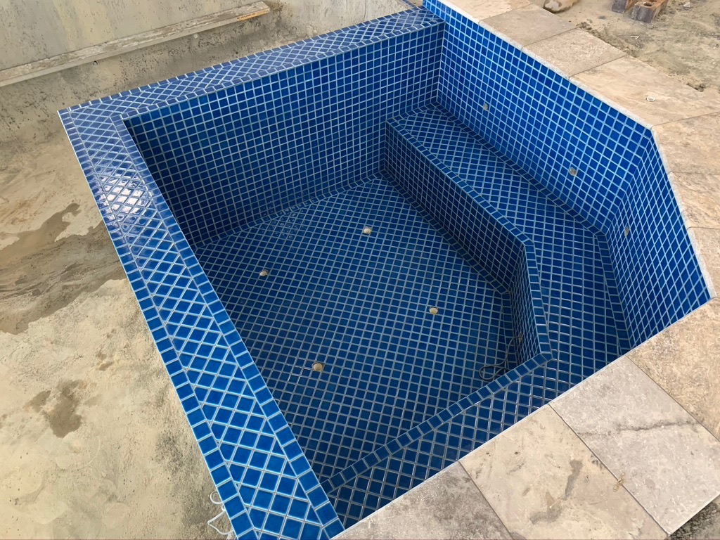 The image showcases a freshly tiled swimming pool that appears to be under construction or repair, featuring a deep blue mosaic tile pattern. The pool has a defined shape with a set of shallow steps leading into it, emphasising both functionality and aesthetics. The surrounding area is unfinished, indicating ongoing work, with concrete flooring visible around the pool's edges.