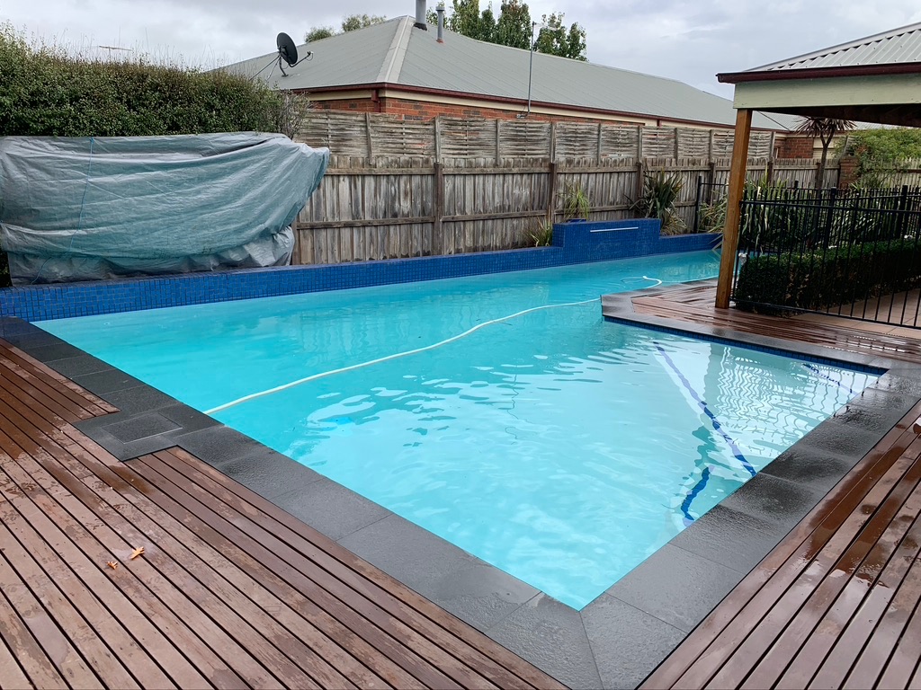 The image showcases a swimming pool featuring clear blue water, surrounded by a wooden deck. The pool appears to be well-maintained, although a section is covered with a grey tarp, indicating either maintenance work or seasonal closing. The backdrop includes a wooden fence and some greenery, contributing to a pleasant outdoor environment.